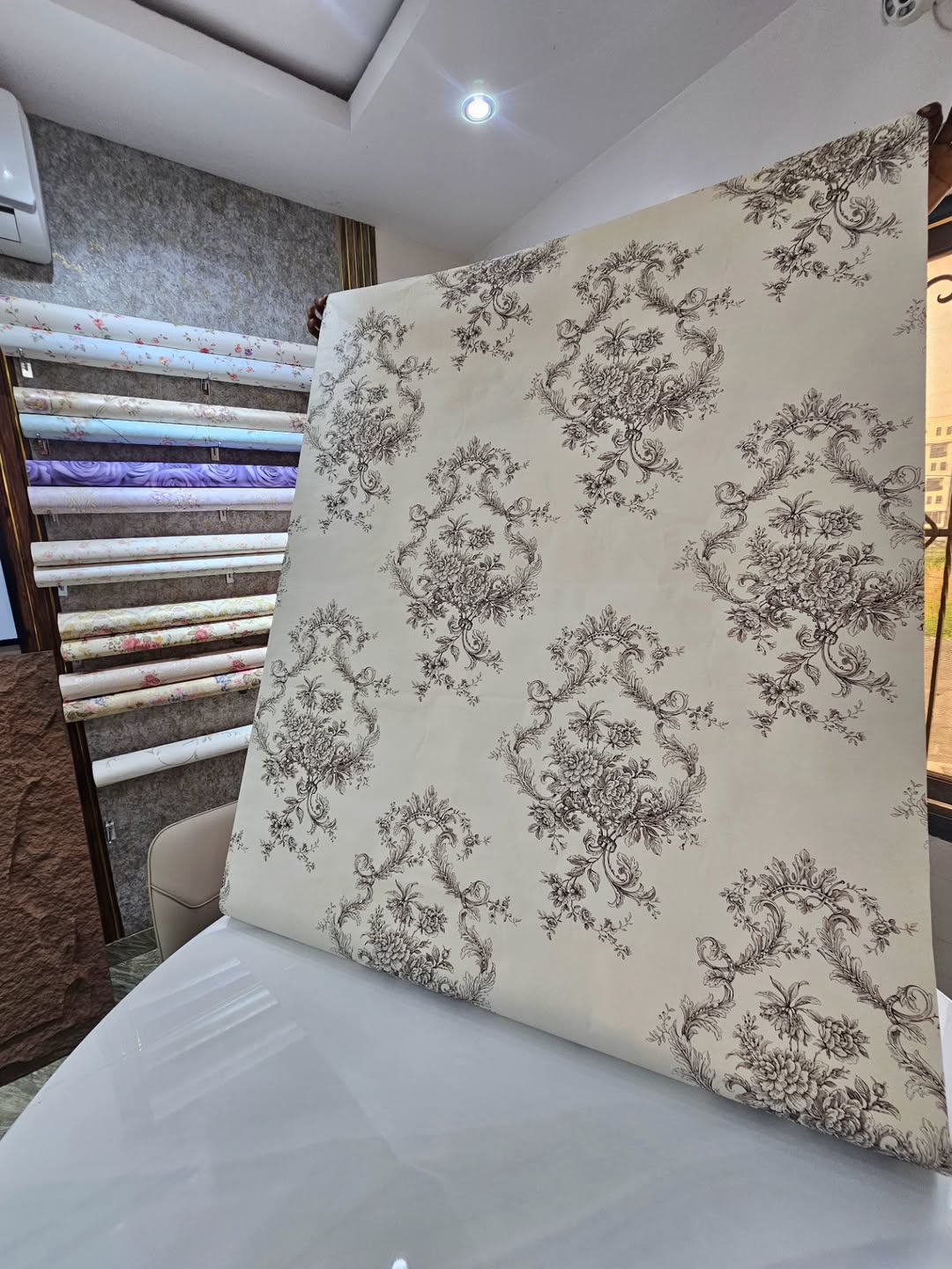 A large sample sheet of off-white wallpaper featuring a repeating monochromatic brown floral medallion pattern. The sheet is held up over a white tabl